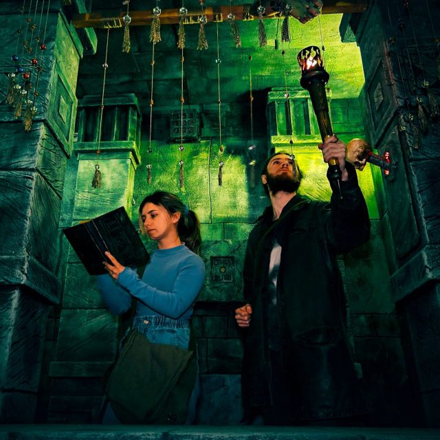 Joueuse tenant un livre et joueur levant une torche dans une salle de pierre verte, salle Tomb Hunter chez Freeing à Saint-Maximin