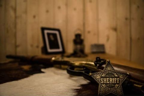 Étoile de shérif et fusil posés sur une peau devant un cadre, mur de planches, salle Evasion au Far-West à Annecy
