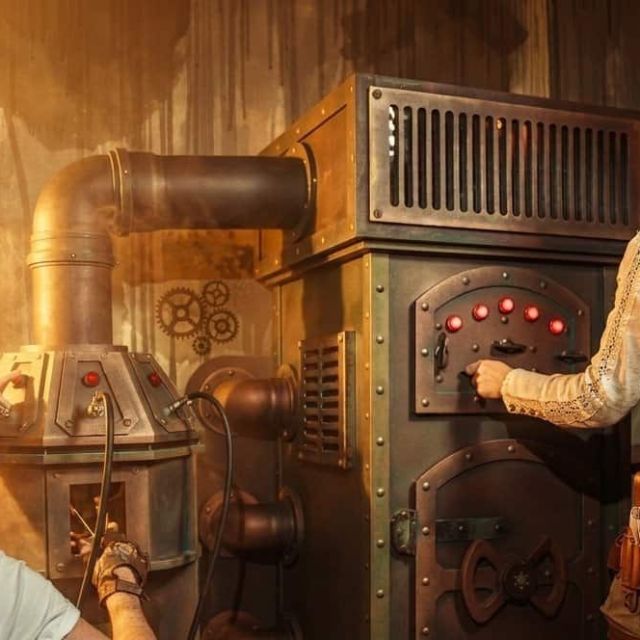 Joueur en chapeau et joueuse devant une machine à tuyaux et boutons rouges, salle Steampunk chez BlackOut Room à Nice
