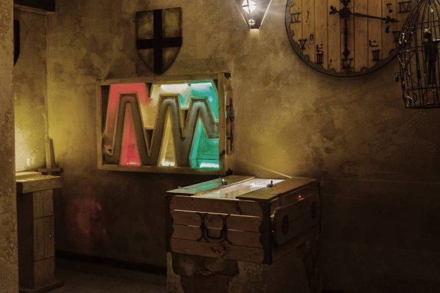 Cachot médiéval aux murs patinés, écran de motifs en M et horloge, salle Les cachots du chevalier noir chez Happy Hour à Paris