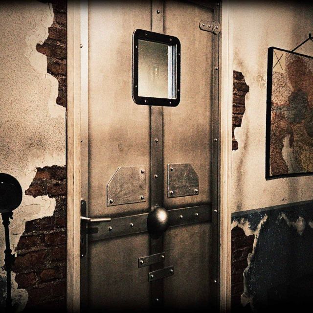 Cellule carcérale décrépite avec porte métallique, miroir et hublot, murs de béton écaillé